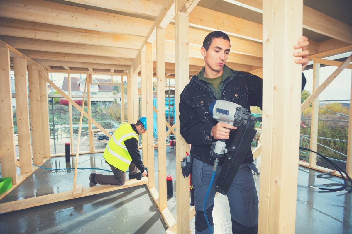 A man on a construction site using a framing nailer to drive nails into a timber frame. 
