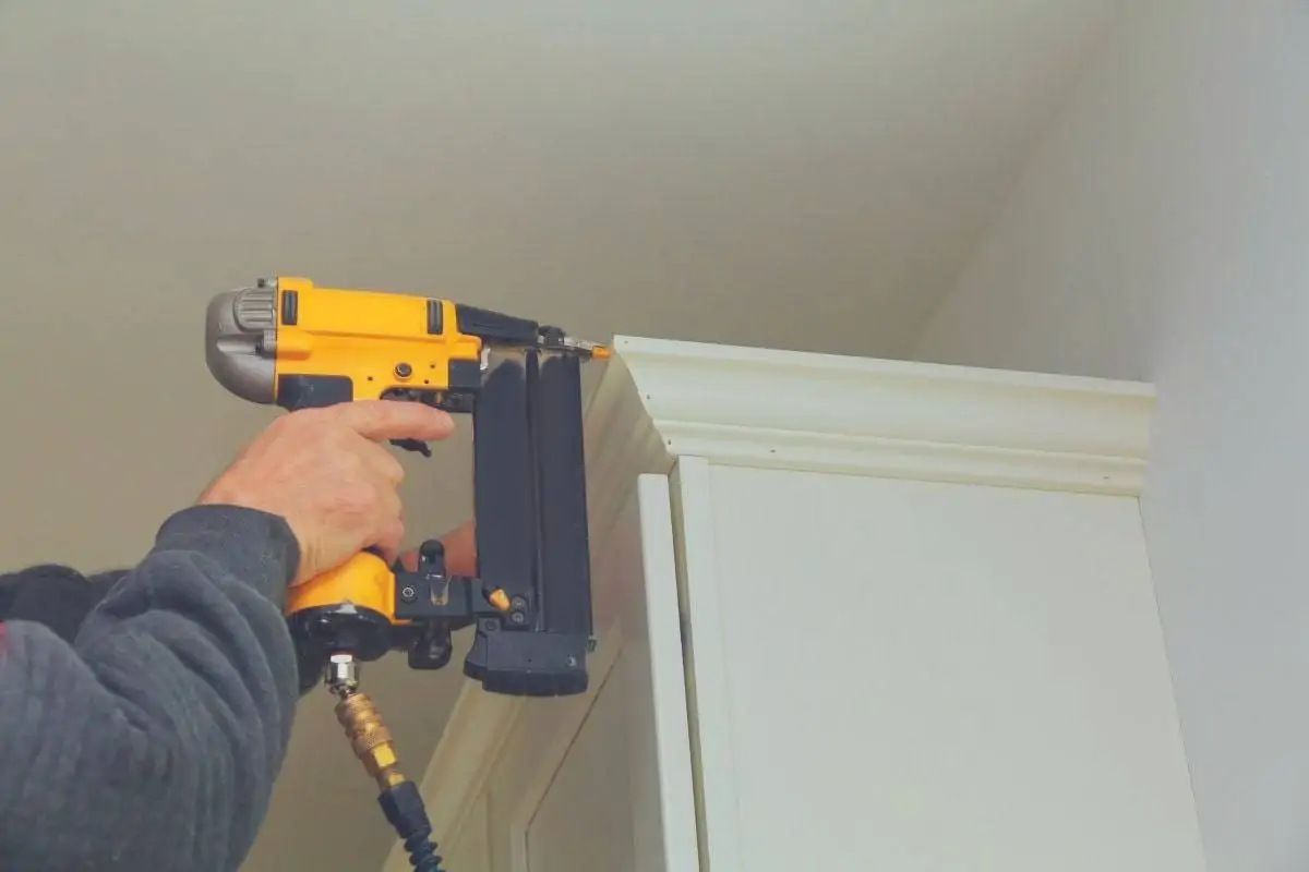 A man using a finishing nail gun to attach some trim to a cupboard.
