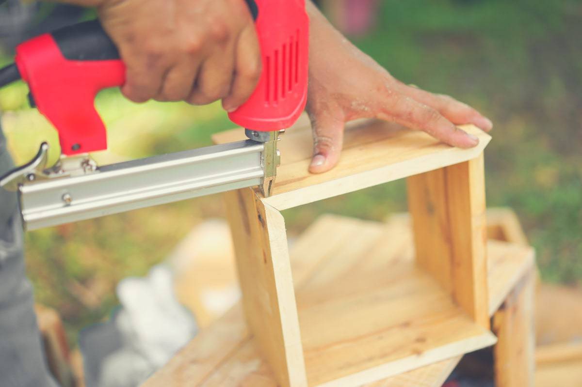 A person using a pin nail gun to fix the corners of a small wooden box

