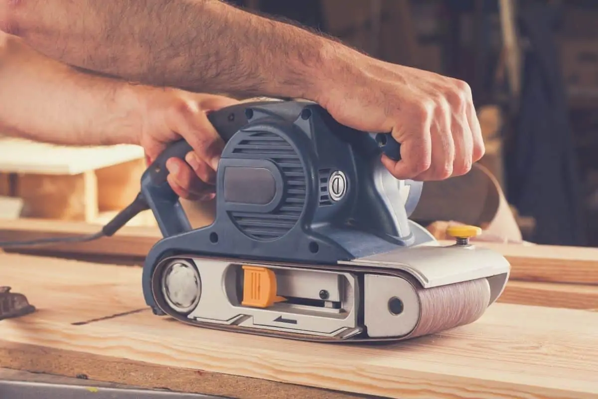A man using a hand-held belt sander to sand a piece of flat wood.
